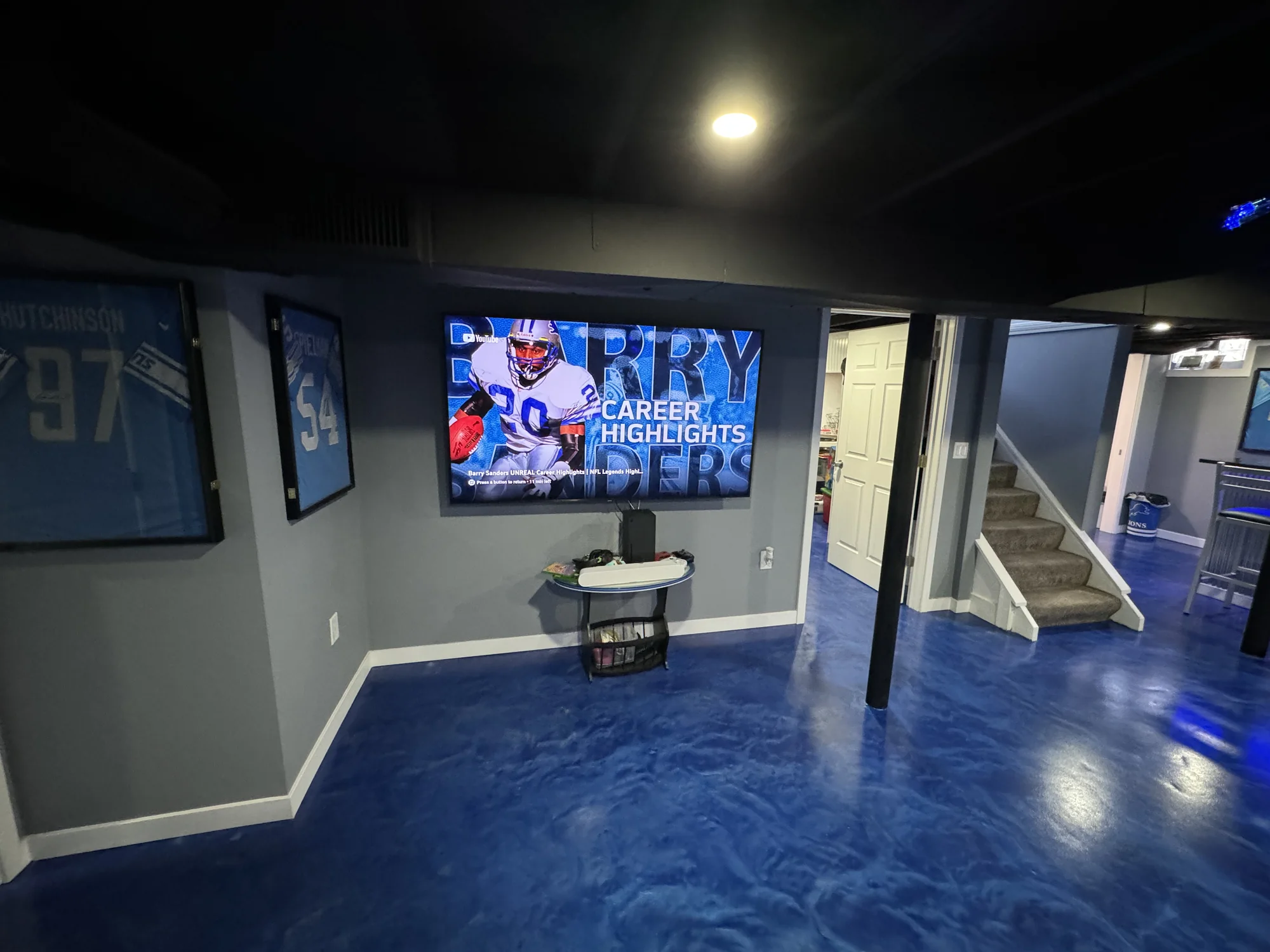 Finished basement theater room with epoxy blue floor, mounted TV showing Barry Sanders highlight, framed Detroit Lions jerseys, and bar area