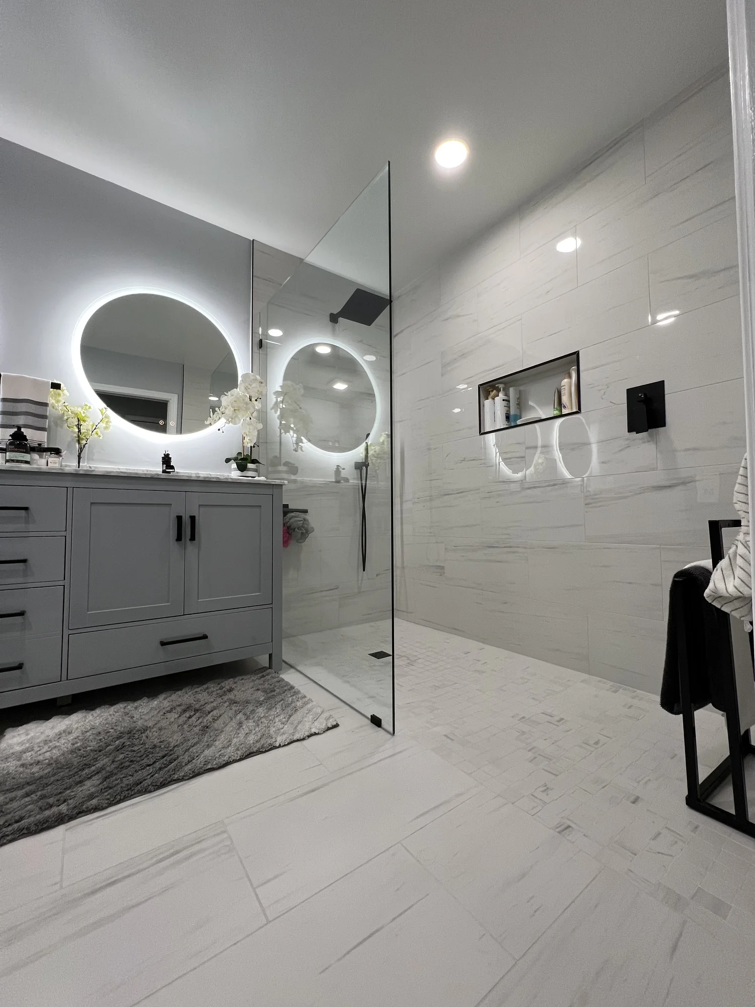 Primary bath with frameless walk-in glass shower, gray marble walls, twin backlit round mirrors, and white shaker vanity