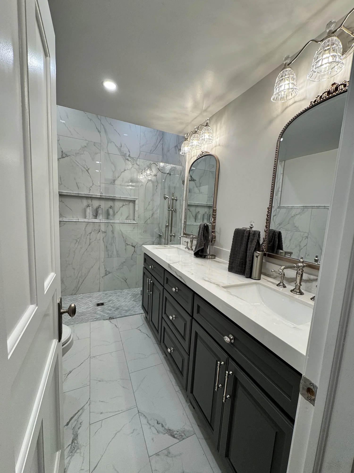 Primary bath with marble tile shower, double black vanity, arched mirrors, and chrome fixtures