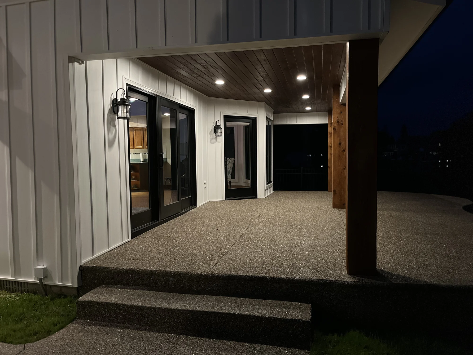 Covered porch entry of modern farmhouse remodel at night — board-and-batten siding, wood columns, brick base, lit pendant overhead