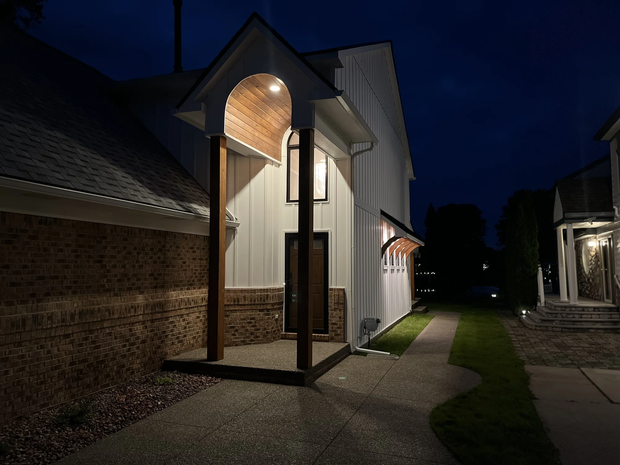 Side angle of modern farmhouse at twilight with lit arched entry, brick base, board-and-batten siding, and wood columns