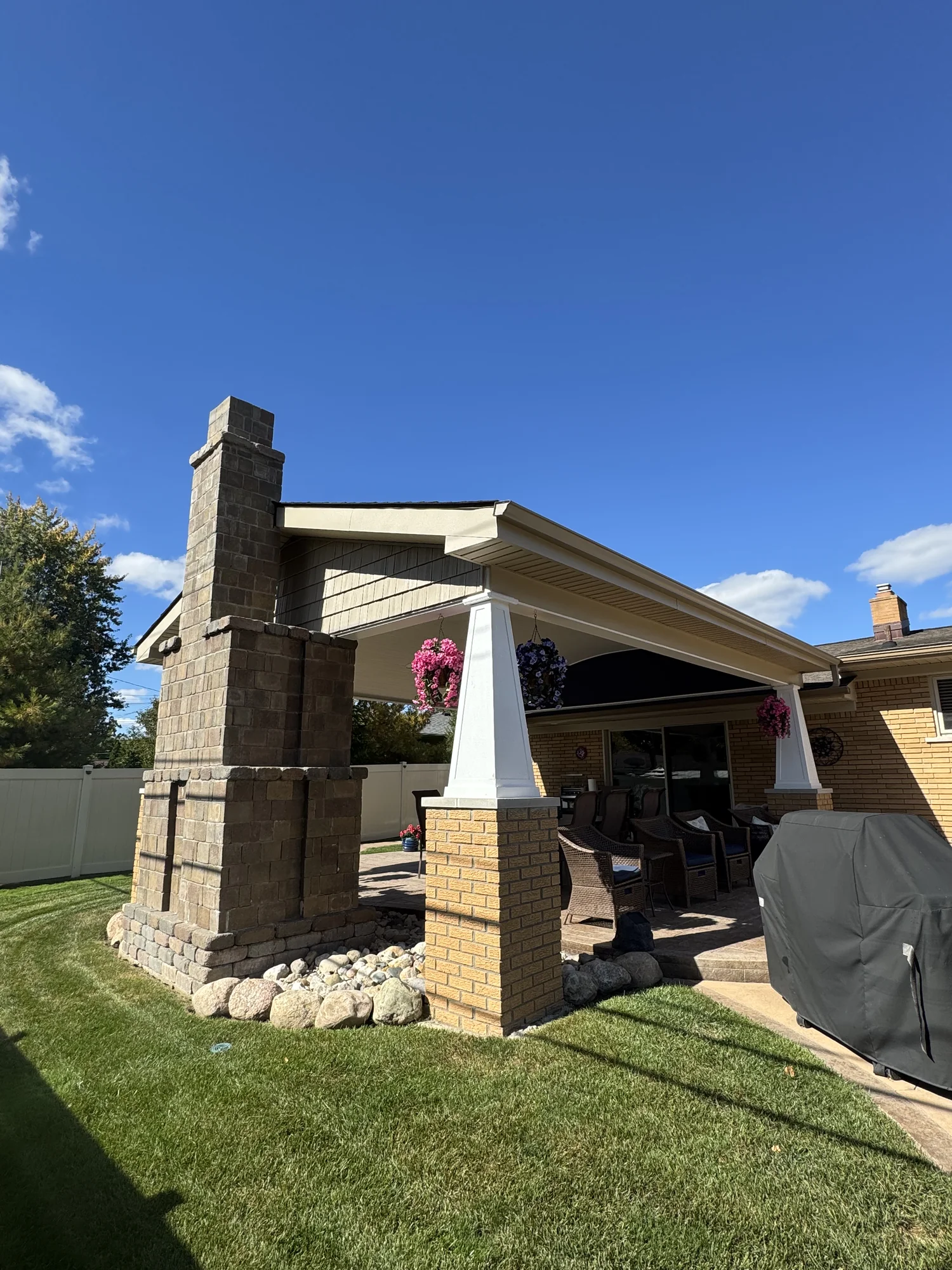 Covered patio pavilion with prominent stone chimney column, tapered brick posts, brick paver patio, and tan ranch home behind