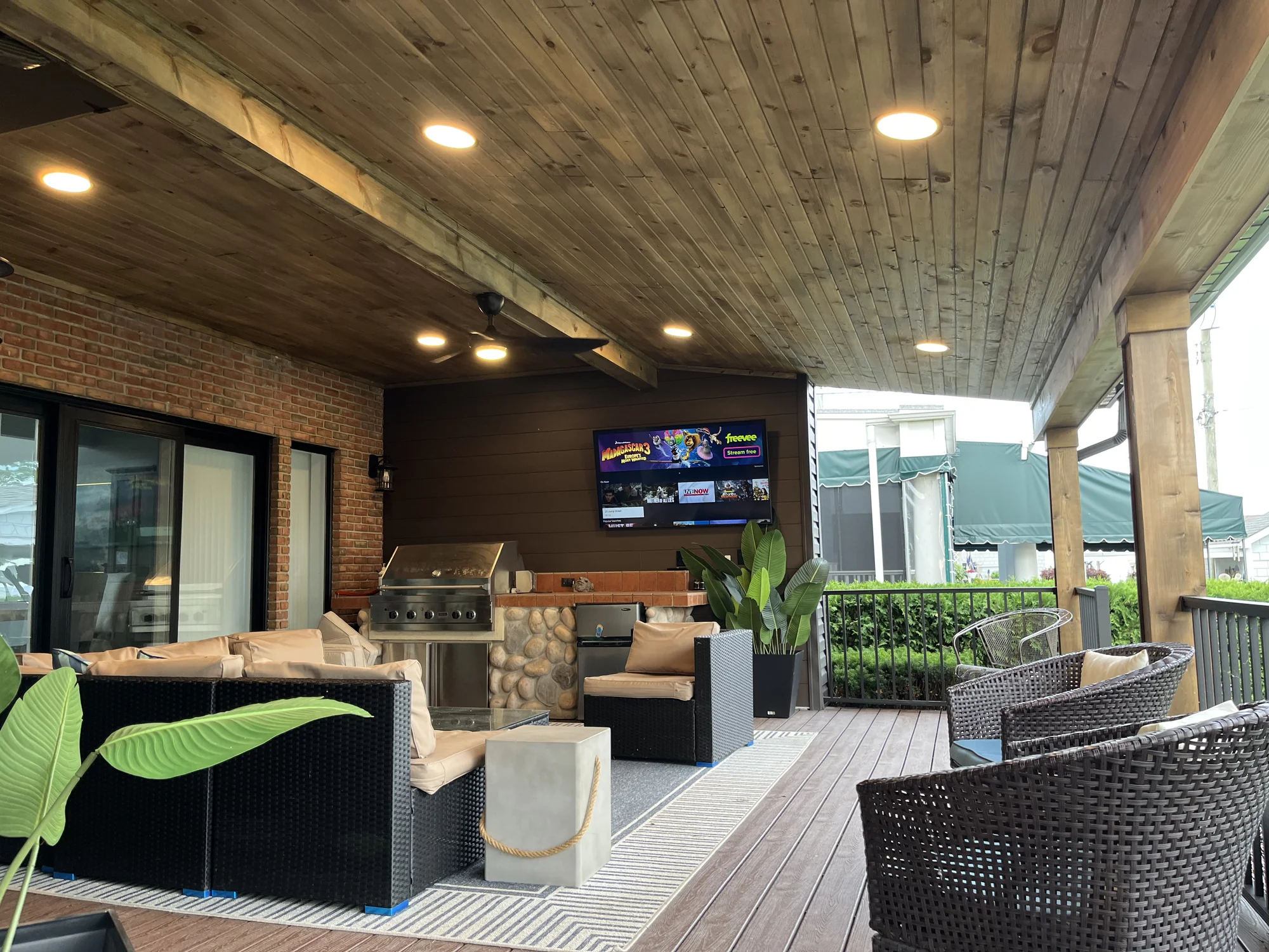 Covered porch with knotty pine ceiling and beam, built-in grill with stone base, wicker lounge seating, and mounted TV